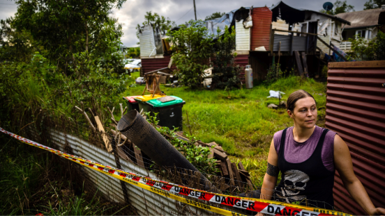 A woman stands before a damaged house. 