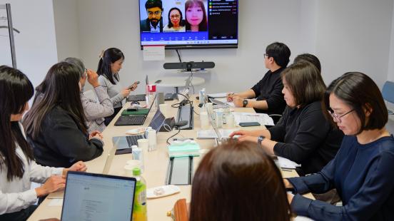 A group of people sit around a conference table with laptops and documents while participating in a hybrid meeting, with several remote participants visible on a wall-mounted video screen ahead.