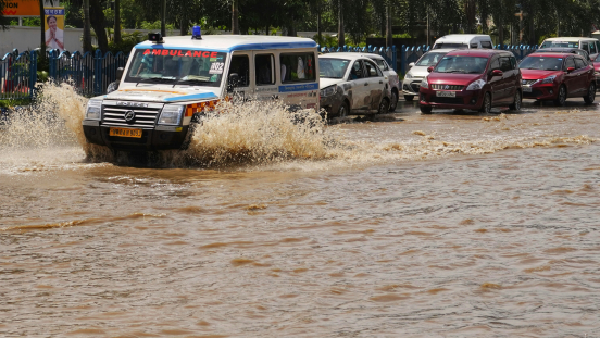 An ambulance drives through a flooded street, followed by other cars.
