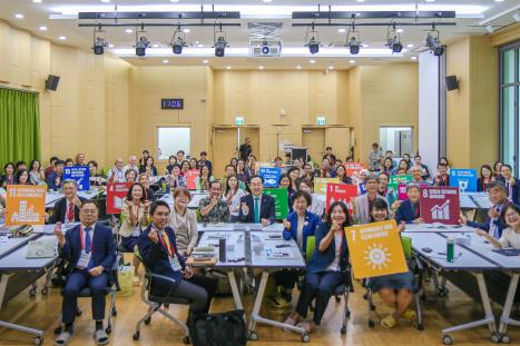 A large group of conference participants seated in a meeting room pose together while holding signs representing different Sustainable Development Goals (SDGs).