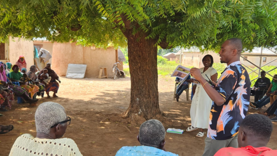 A group of Ghanians sit under a tree in a circle, while a woman and man are standing in the middle talking and showing a book.
