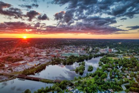 Aerial view of a city at sunset, with a river winding through green spaces beneath clouds.