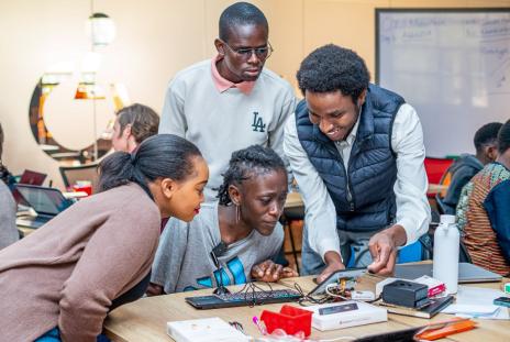 Four people gather around a table in a classroom or workshop space, examining an electronics project with wires, components and a computer keyboard.
