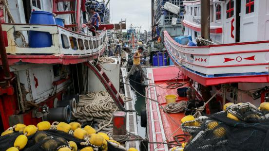 Several workers aboard multiple large fishing ships docked in Thailand.