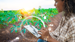 A female farmer next to a farm looking at data on her tablet device, with environment-related icons overlayed on top