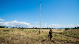 A man stands next to an iron tower in a field. 