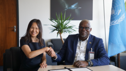 Two persons smiling and shaking hands after signing a document in front of a UN flag