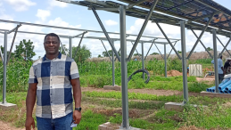 A man poses in an agricultural field underneath a solar panel tower. 