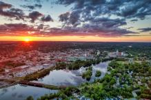 Aerial view of a city at sunset, with a river winding through green spaces beneath clouds.