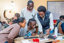 Four people gather around a table in a classroom or workshop space, examining an electronics project with wires, components and a computer keyboard.