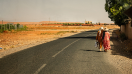 Two women with hats walk with their back towards us on a road surrounded by dryland. 