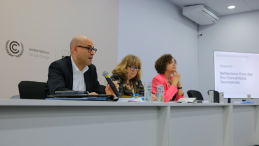 A man and two women sit at a panel desk in a conference setting