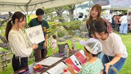 A child participates in an educational activity at an outdoor community event, interacting with facilitators under a tent, with illustrated materials displayed on a table.