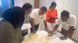 Participants collaborate around a table during a workshop activity, reviewing cards and worksheets while taking notes.