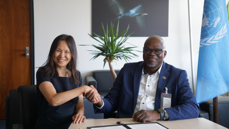 Two persons smiling and shaking hands after signing a document in front of a UN flag