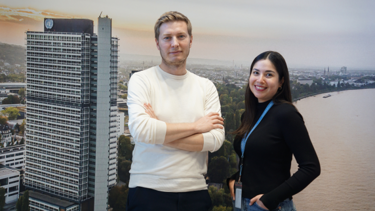 Two people standing in front of a background of a UN building at the river Rhine. 