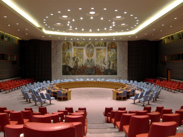 UN Security Council meeting space with circle of chairs and one connected desk facing each other for members and red audience seats surrounding with gold and colored mosaic on back wall