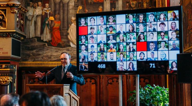 UNU-CRIS event with Philippe De Lombaerde standing at a podium with a screen behind him displaying profile pictures of UNU-CRIS personnel