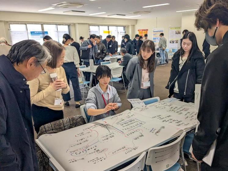 A group of participants stand around a table covered with handwritten notes and diagrams as one person presents during a collaborative workshop.