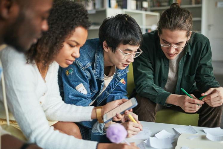 Four people lean over a table covered with papers and notes, closely collaborating as they write and look at a smartphone in a bright indoor workspace.