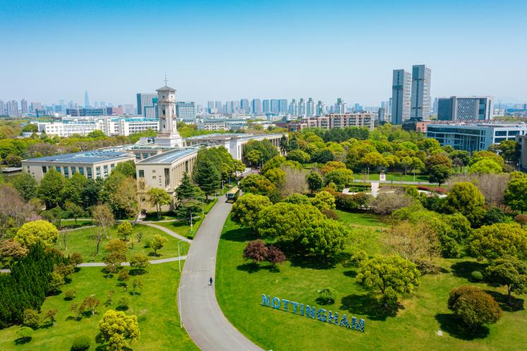 A university clock‑tower building sits at the center of a green, tree‑filled campus with “NOTTINGHAM” spelled out in large blue letters on the lawn and a modern city skyline rising behind it.