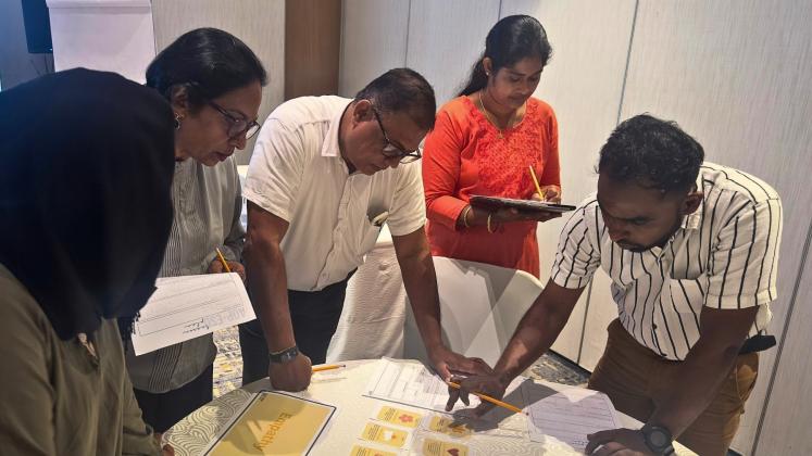 Participants collaborate around a table during a workshop activity, reviewing cards and worksheets while taking notes.