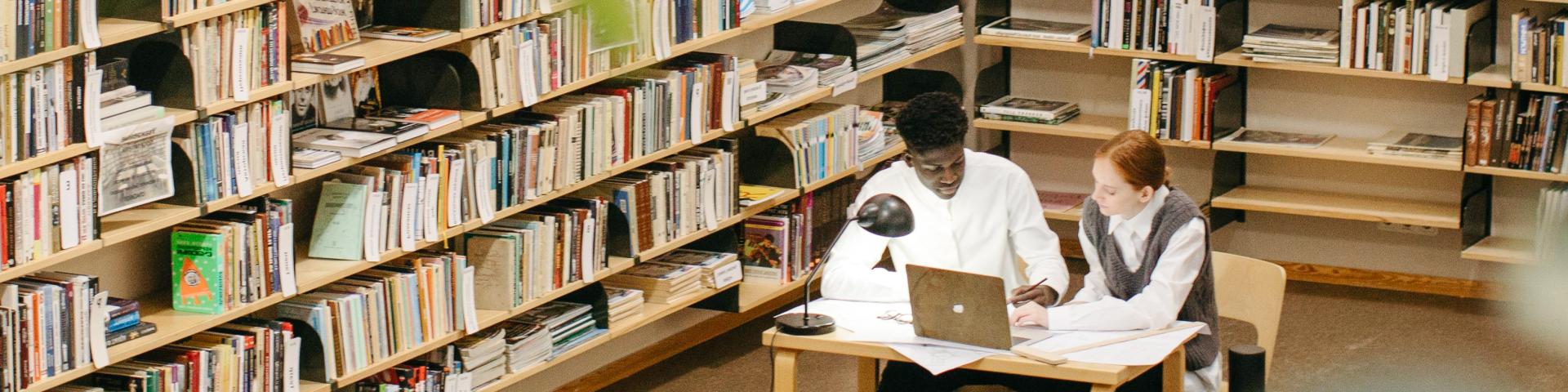 Two people work together at a table with a laptop in a library, surrounded by tall shelves filled with books and study materials.